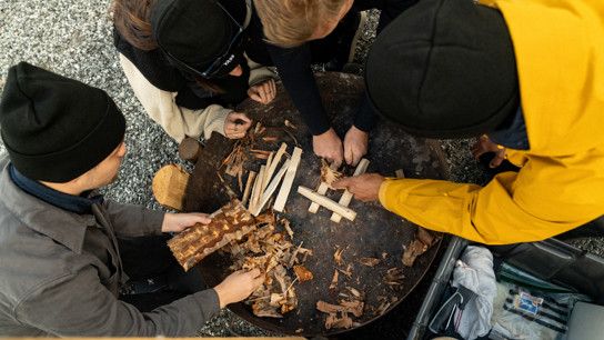 Flera människor i friluftskläder samarbetar kring en grillplats i fjällmiljö 