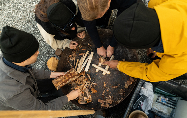 Flera människor i friluftskläder samarbetar kring en grillplats i fjällmiljö 
