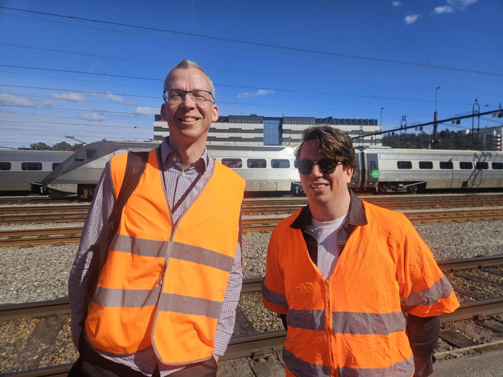 Två glada män som jobbar med test står framför ett tåg en solig dag