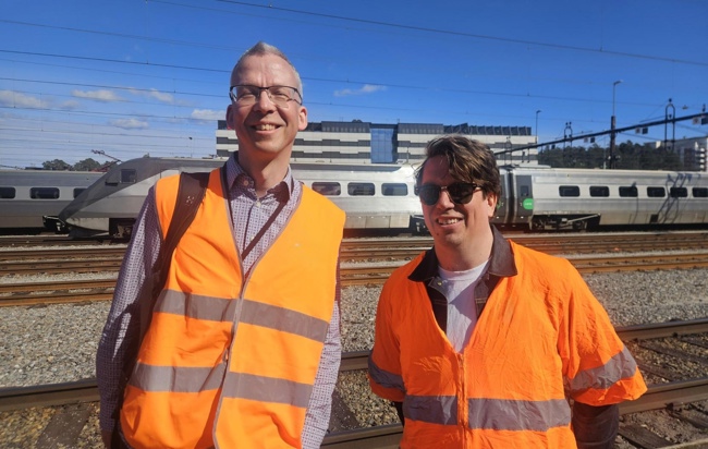 Två glada män som jobbar med test står framför ett tåg en solig dag