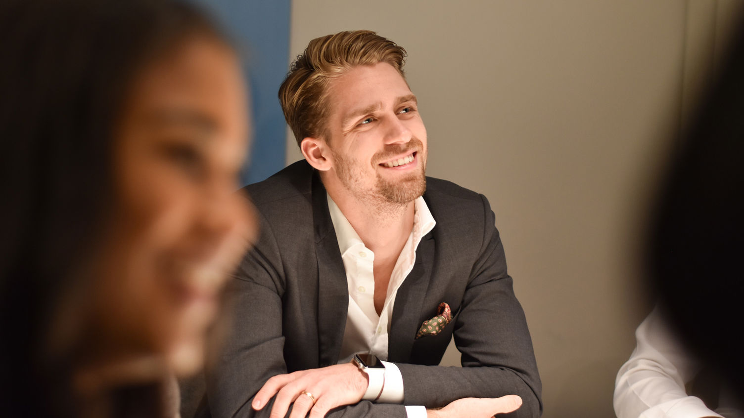 En leende man i grå kostym och vit skjorta sitter vid ett bord och tycks lyssna på någon prata.