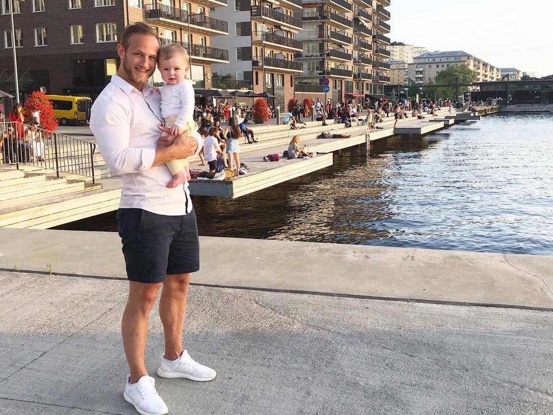 En man står med ett barn i famnen framför en kaj, omgiven av stadsmiljöns liv och rörelse.